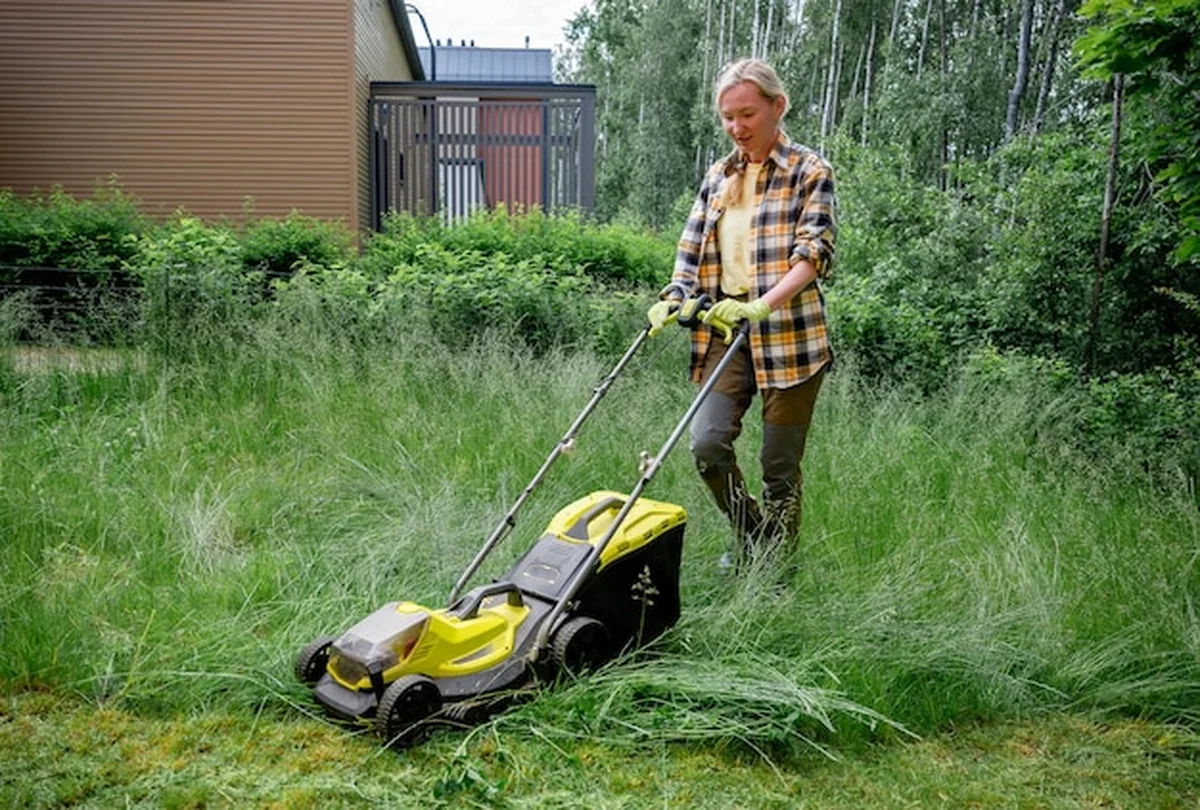 Kobieta używa elektrycznej kosiarki w zarośniętym ogrodzie obok drewnianego domu